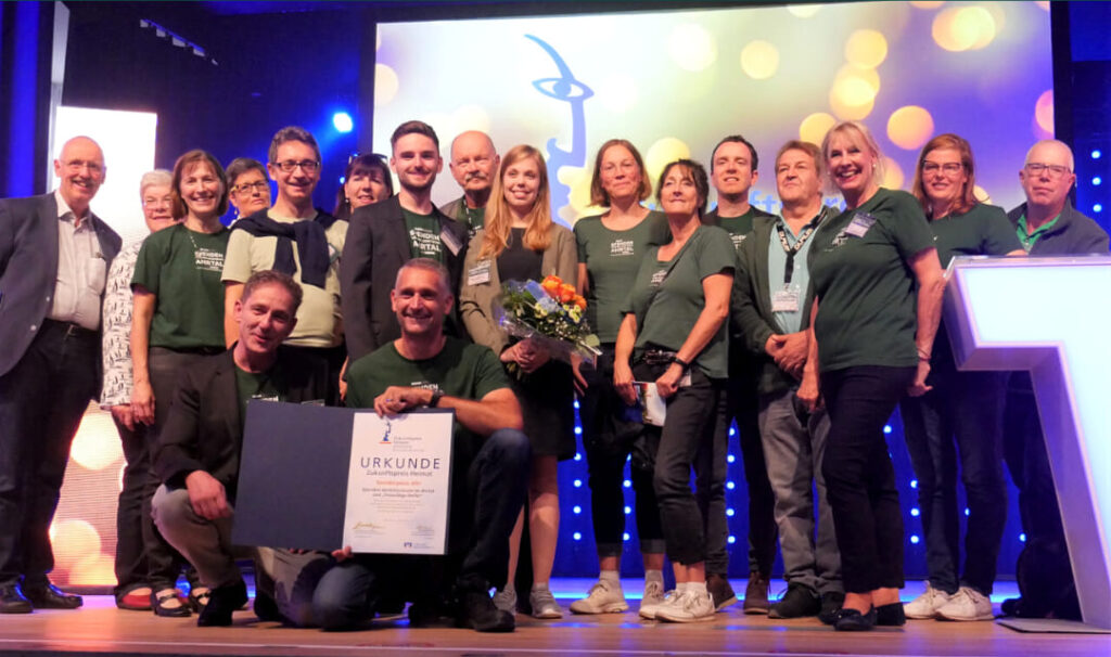Das Team des Spenden-Verteilzentrums Ahrtal steht gemeinsam für ein Gruppenfoto auf der Bühne mit einer Urkunde des Zukunftpreis Heimat.