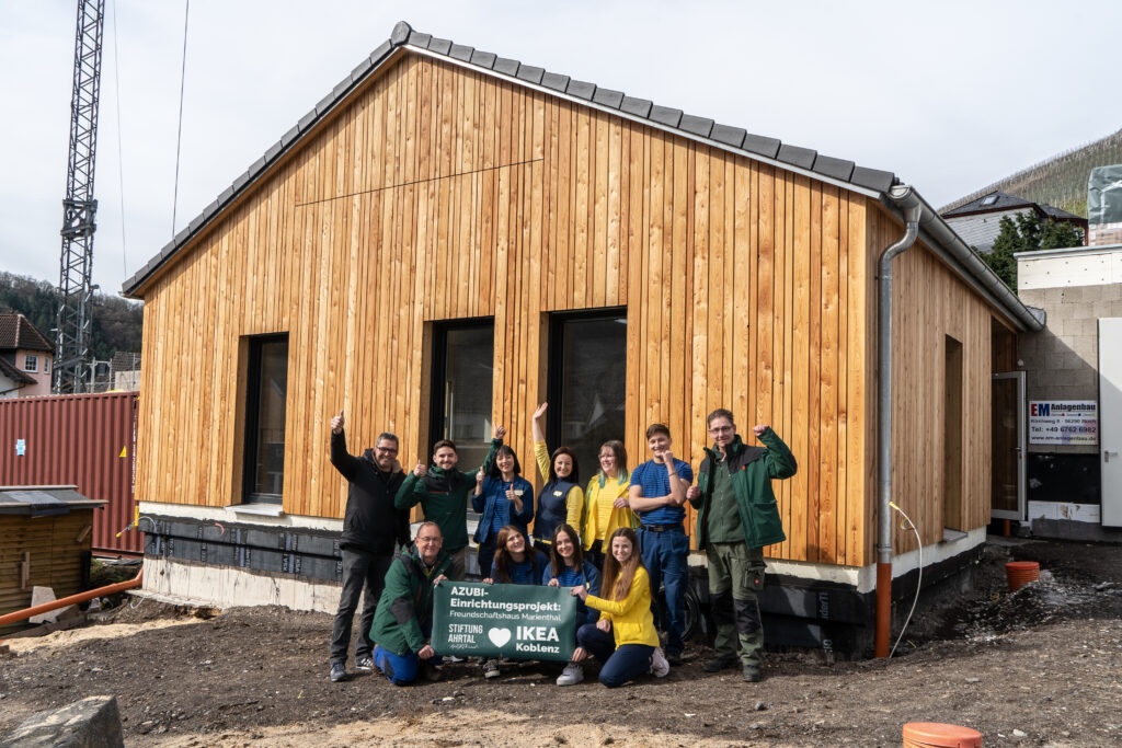 Eine Gruppe aus IKEA Mitarbeiten und Helfern des Spendenverteilzentrums jubelt vor dem Freundschaftshaus in Marienthal. Dabei halten sie einen Banner ihres Kooperationsprojektes hoch.